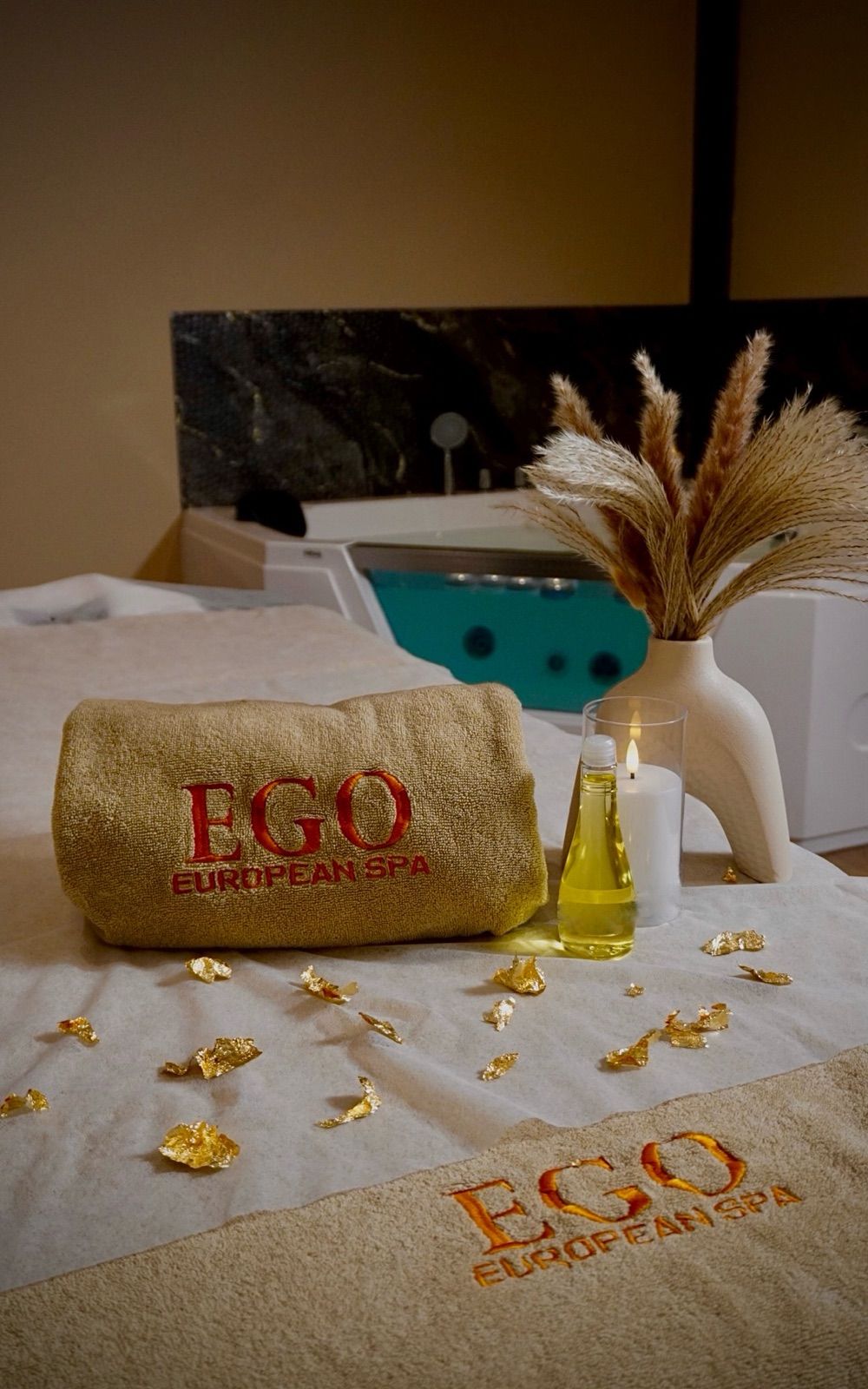 Treatment bed with a rolled EGO European Spa towel, massage oil, a lit candle and gold flakes, with the filled jacuzzi and pampas grass behind it