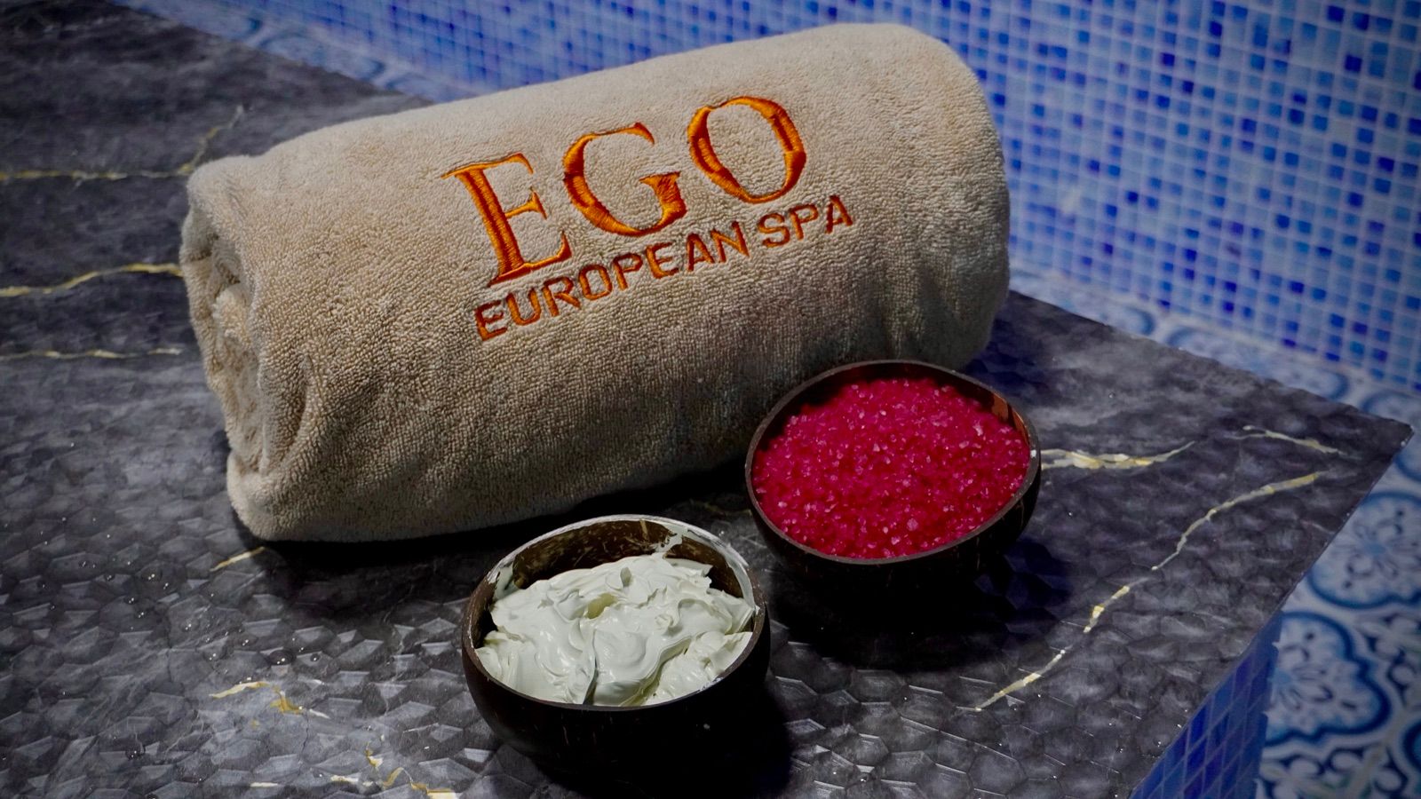 Rolled EGO European Spa towel beside coconut-shell bowls of bath salt crystals and body cream, on dark marble at the mosaic jacuzzi in Deira, Dubai
